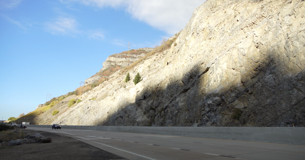 Looking at U.S. Route 189 in Provo Canyon and the newly completed barrier, with sunny skies, harsh shadows from the canyon wall, and beautiful fall colors in the canyon.